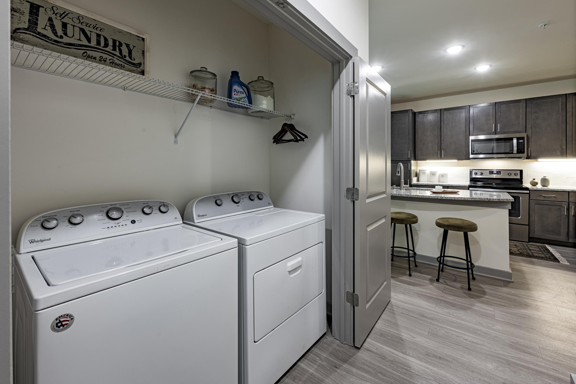 Modern kitchen and laundry area with washer, dryer, shelves, and view of countertop with stools.