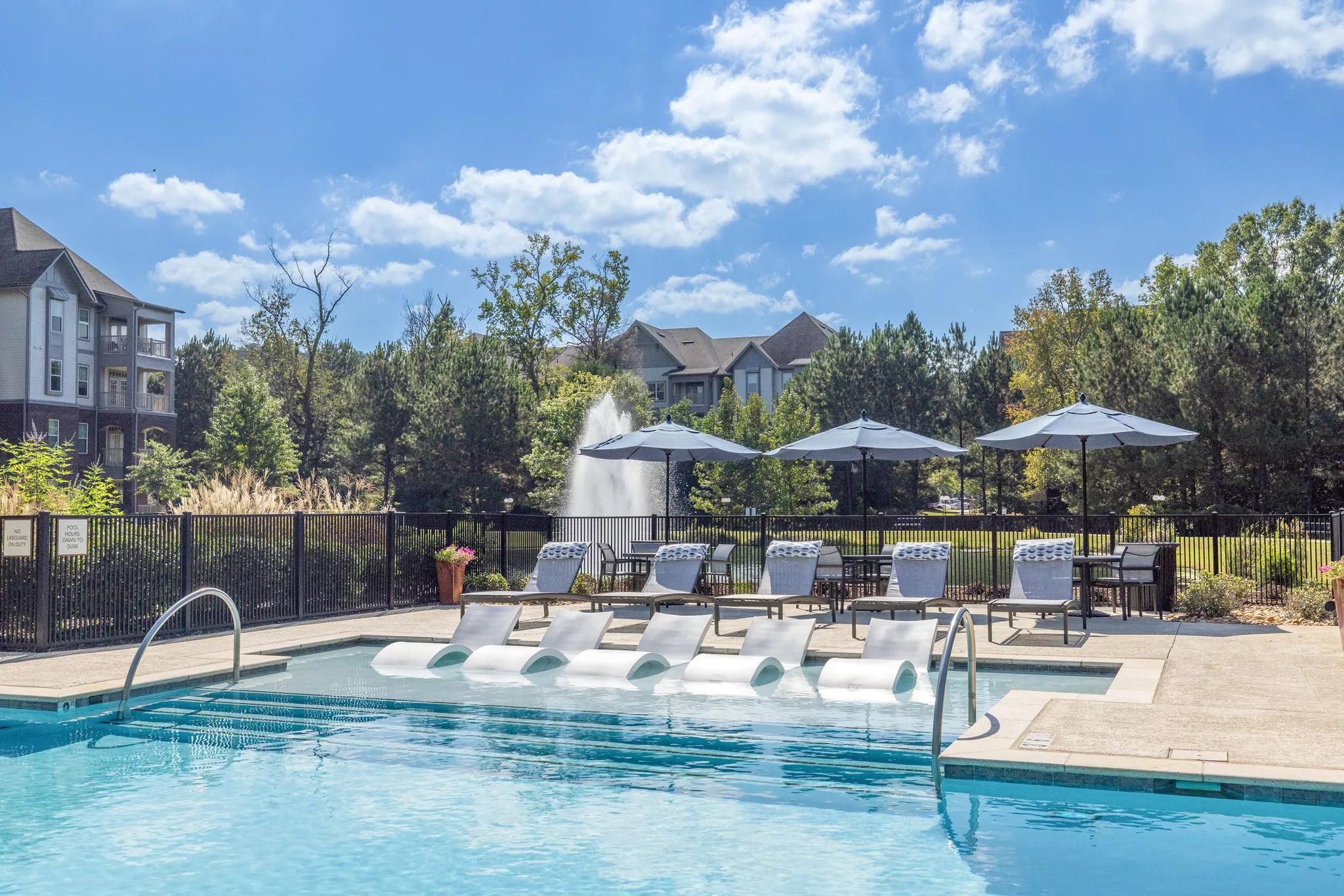 Outdoor pool with lounge chairs, umbrellas, and a fountain, surrounded by trees and apartment buildings.