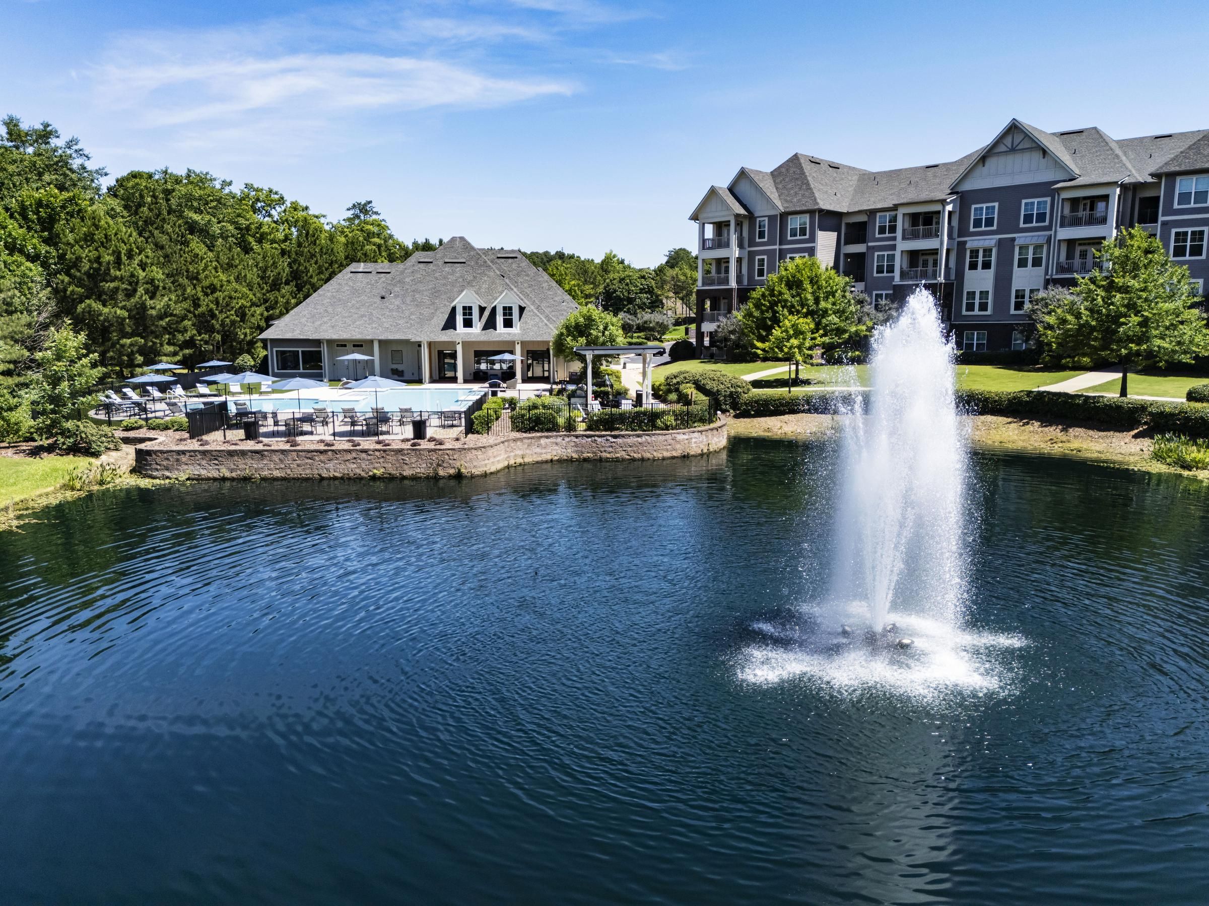 Apartment building and clubhouse by a pond with a water fountain, trees, and outdoor seating areas.