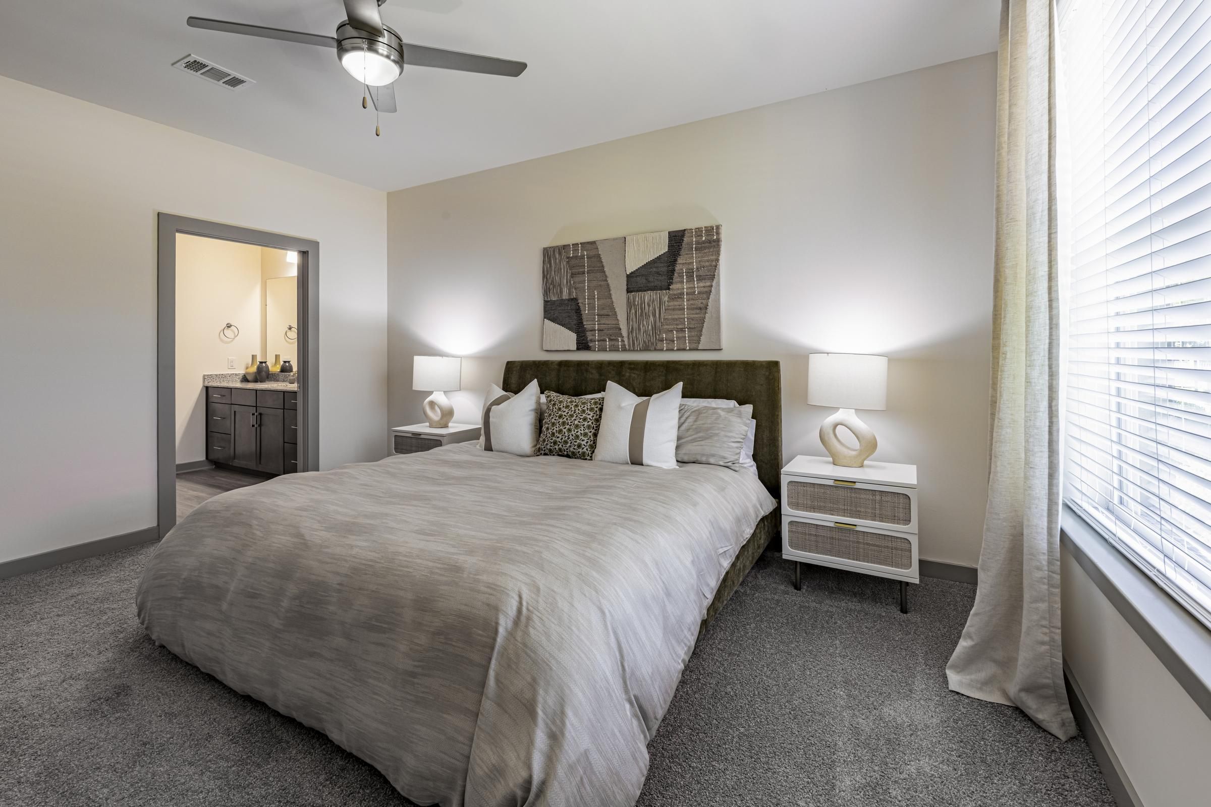 Modern bedroom with a large bed, two nightstands, lamps, wall art, and sunlight streaming through a window.
