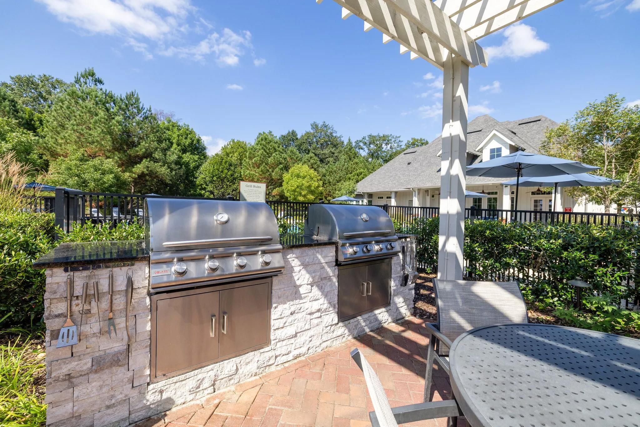 Two outdoor stainless steel grills on a stone patio with trees and a house in the background.
