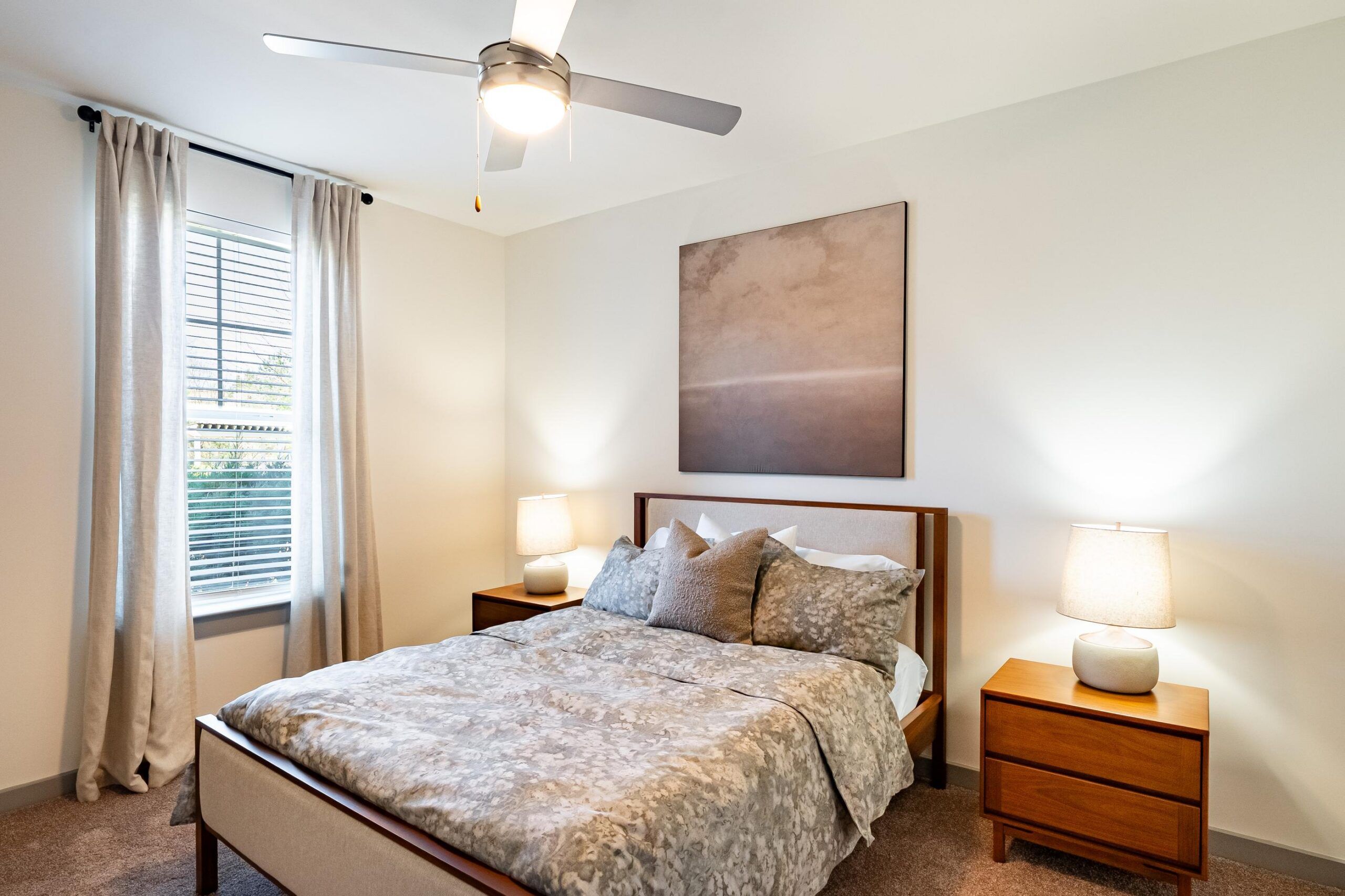 Modern bedroom with a double bed, two nightstands, lamps, artwork above the bed, and window with curtains.