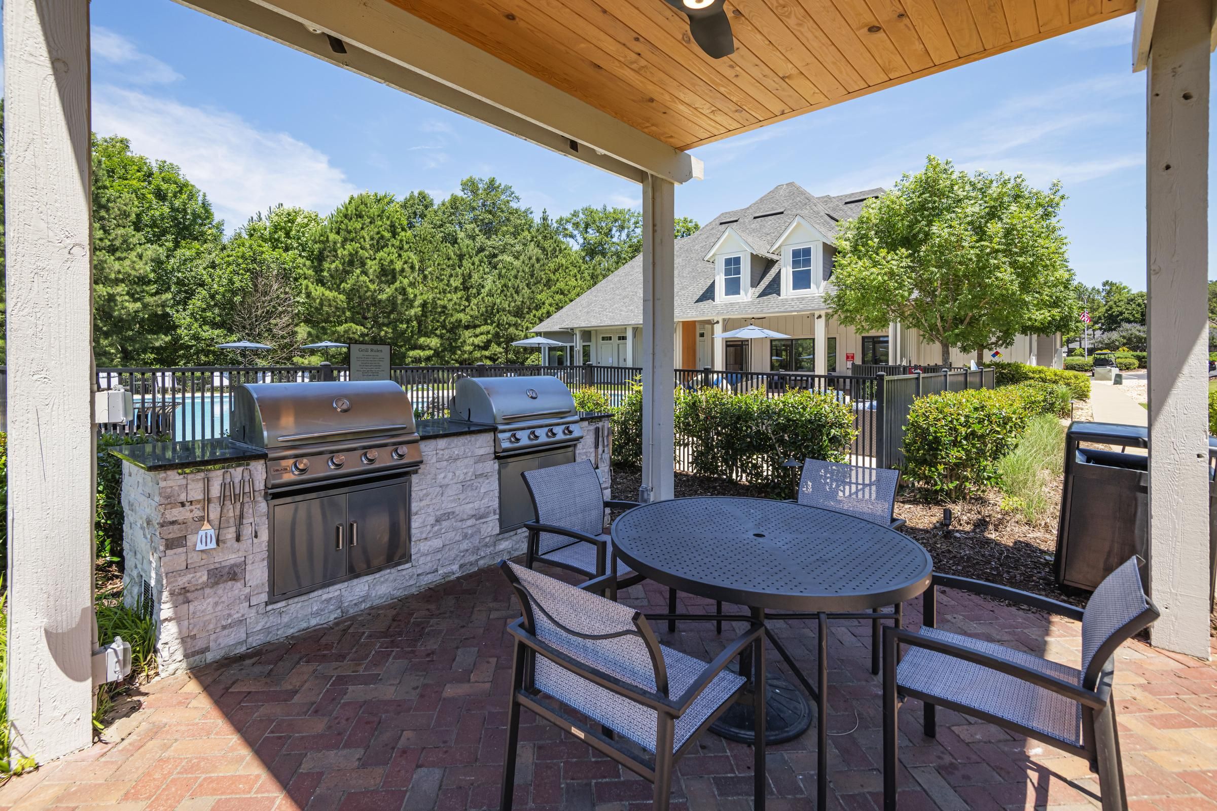 Outdoor patio with two grills, a round table, four chairs, and a view of a house and trees in the background.
