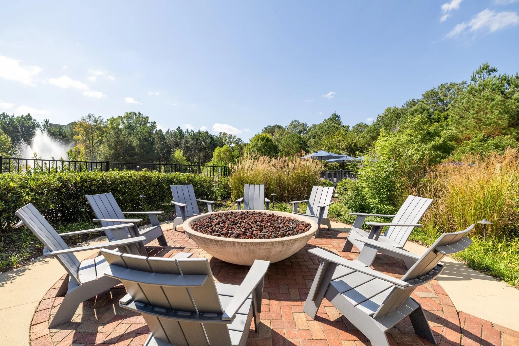 Gray Adirondack chairs surround a circular fire pit on a brick patio, with greenery and blue sky in the background.
