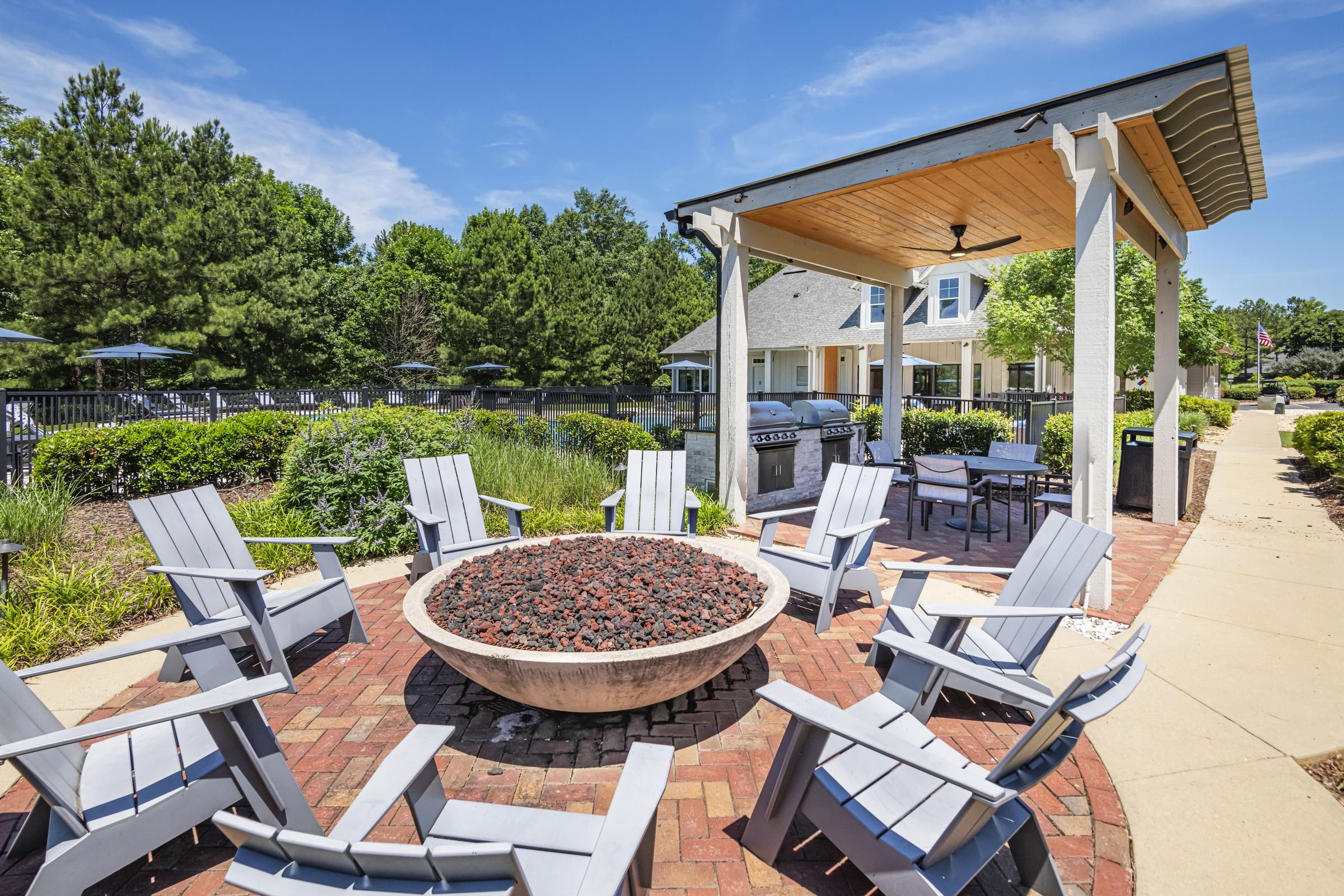 Outdoor patio with white chairs around a fire pit, pergola, and green trees in the background.